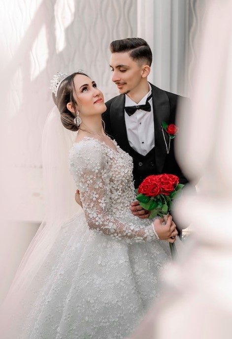 A groom in a black tuxedo and a bride in a lace gown with a veil holding red roses