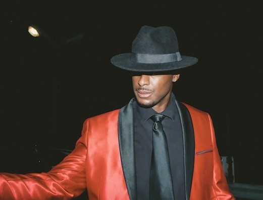 A man in a red satin tuxedo jacket with a black shirt, black tie and a black fedora