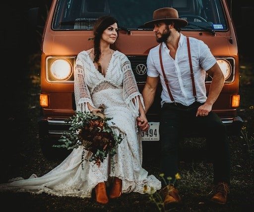 A bride in a lace boho gown and a groom in suspenders and a brown hat sitting on the front of an orange camper van