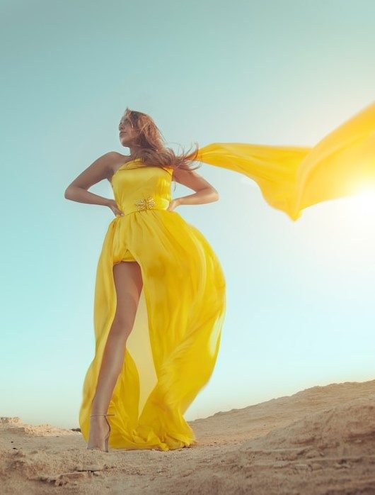 A woman in a flowing yellow gown with the skirt and scarf blowing in the wind