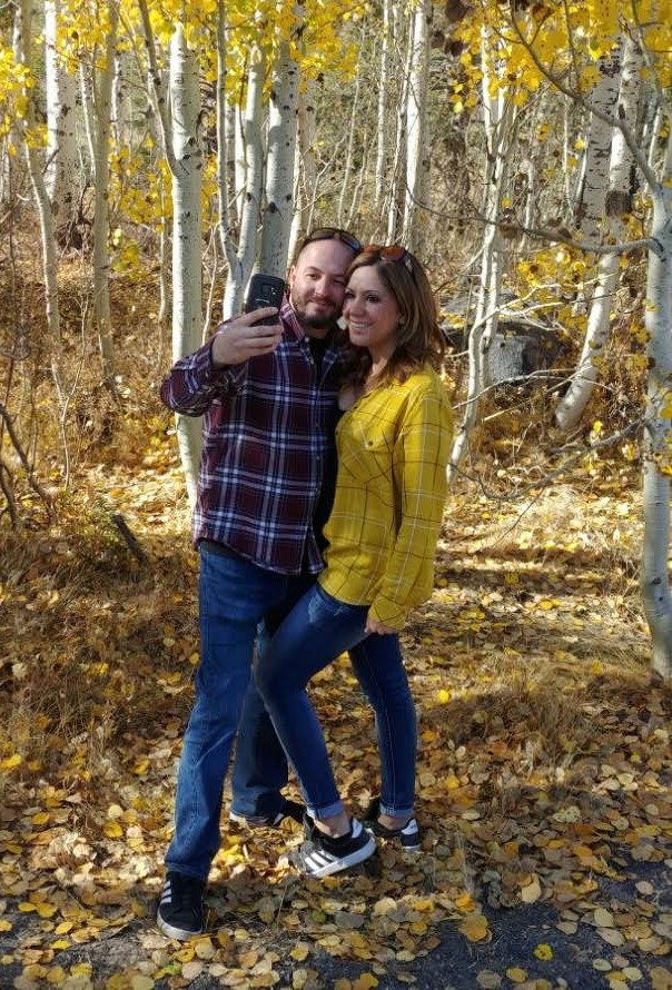 A couple taking a selfie among yellow aspen trees, he in a plaid flannel and she in a mustard yellow top