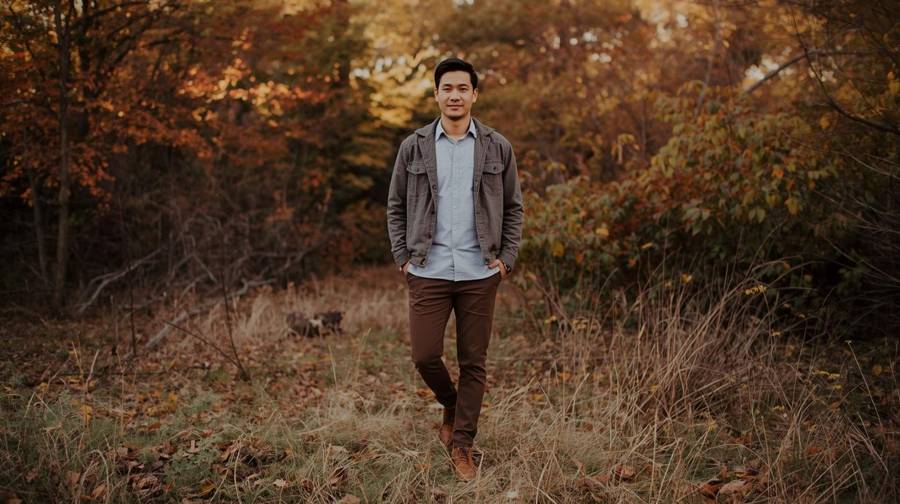 A man in a gray jacket over a light blue shirt, brown pants and boots walking through an autumn woodland path