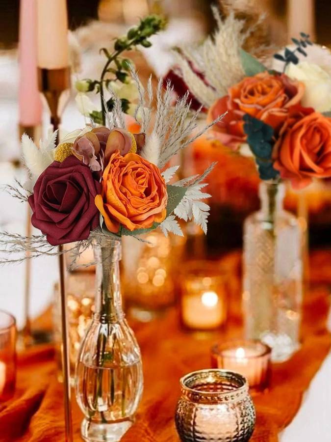 Burgundy and burnt-orange roses with pampas grass in glass bud vases among candles on a terracotta runner