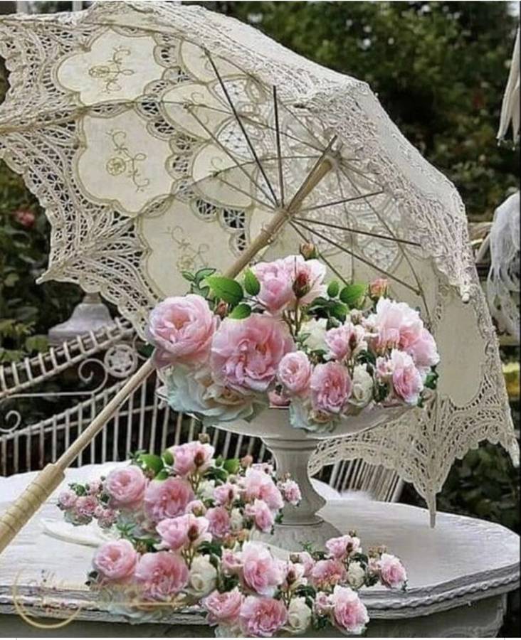 A cream lace parasol resting over a bowl of pink roses on a garden table