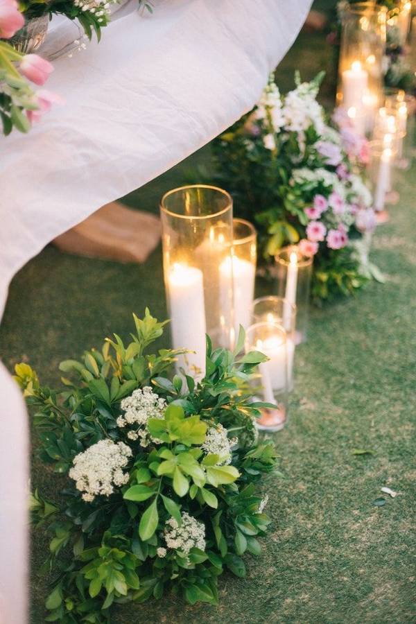 Tall candles in glass cylinders beside greenery and white and pink flowers along the grass