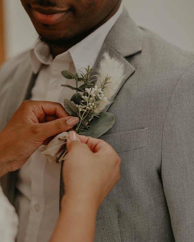 A boutonniere of greenery, small white flowers and a feather being pinned to a gray suit jacket