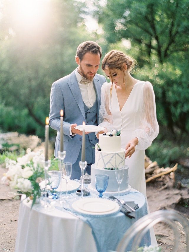 A couple cutting a tiered blue and white cake beside a candlelit table outdoors