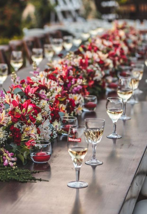 A long wooden table with a runner of red, pink and white flowers and glasses of white wine