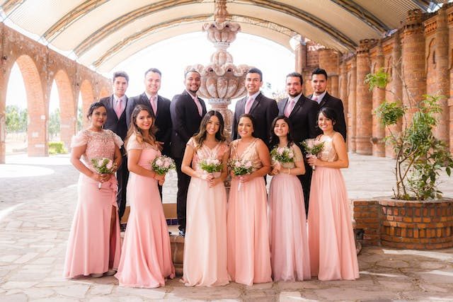 A wedding party of bridesmaids in blush pink dresses and groomsmen in dark suits standing together in a stone courtyard