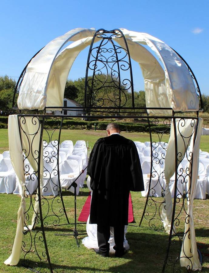 An officiant in a black robe facing rows of white chairs beneath a draped wedding arch