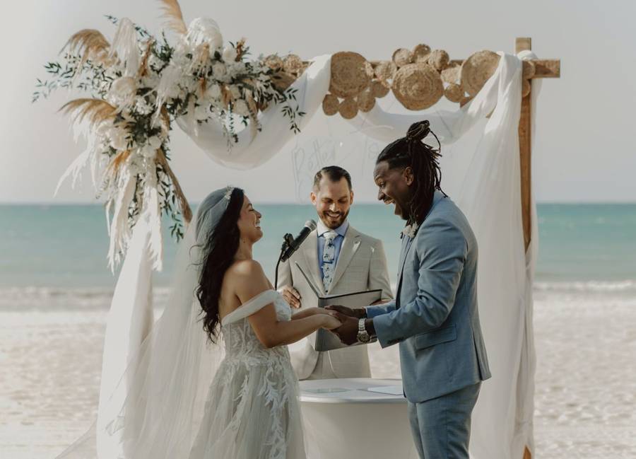 A smiling officiant with a microphone leading a couple through their vows on the beach