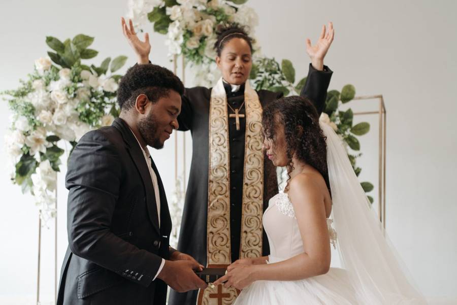 A clergy officiant in a black robe and gold stole raising her hands over a couple holding a Bible