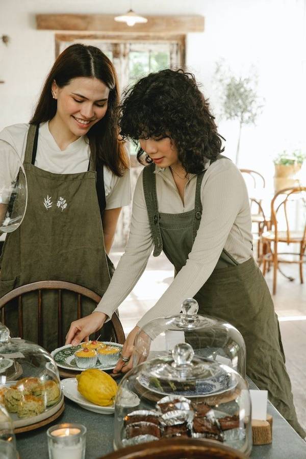 Two servers in aprons setting out cupcakes and brownies under glass domes