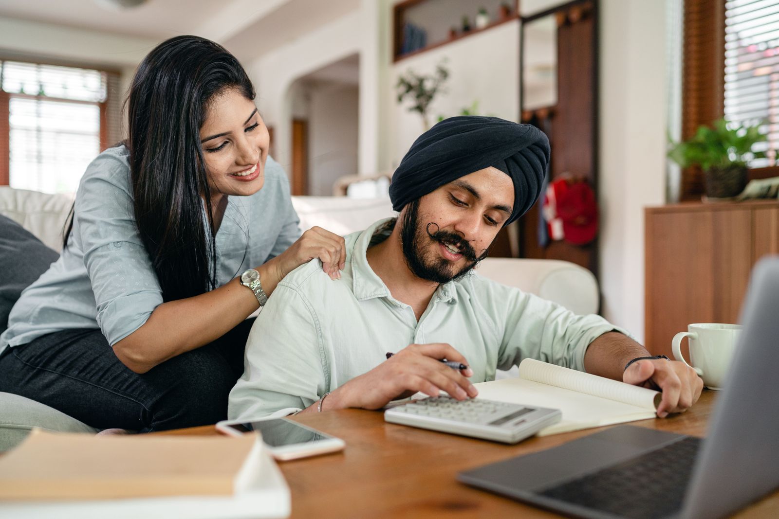 A smiling couple working through their wedding budget together at the kitchen table with a calculator and notebook