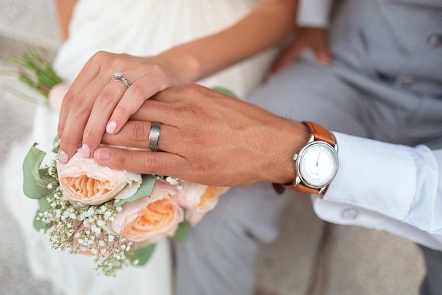 A bride's hand with an engagement ring resting on a groom's hand wearing a plain band and a leather-strap watch