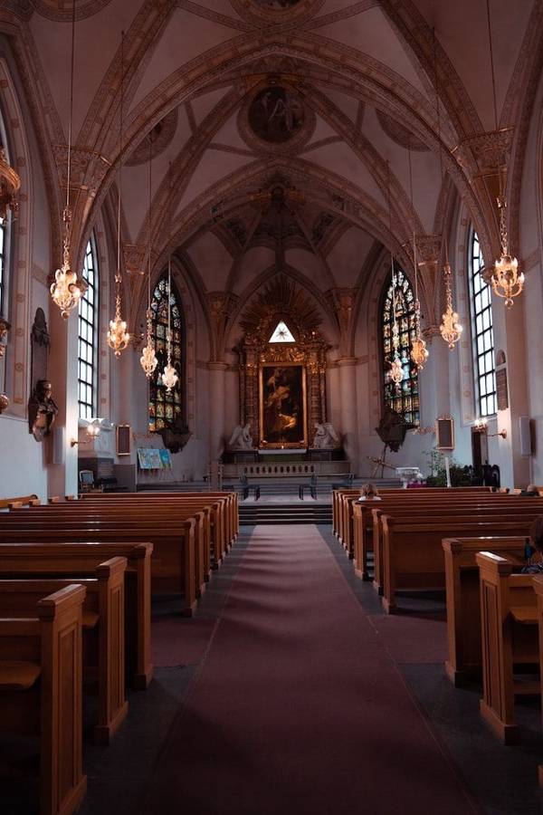 The aisle of a church with wooden pews, chandeliers and stained glass windows