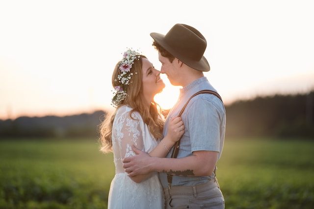 A couple in a green field at sunset, she in a flower crown and he in a brimmed hat and suspenders