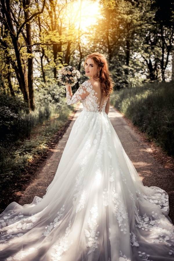 A bride in a lace gown with a long train on a sunlit forest path