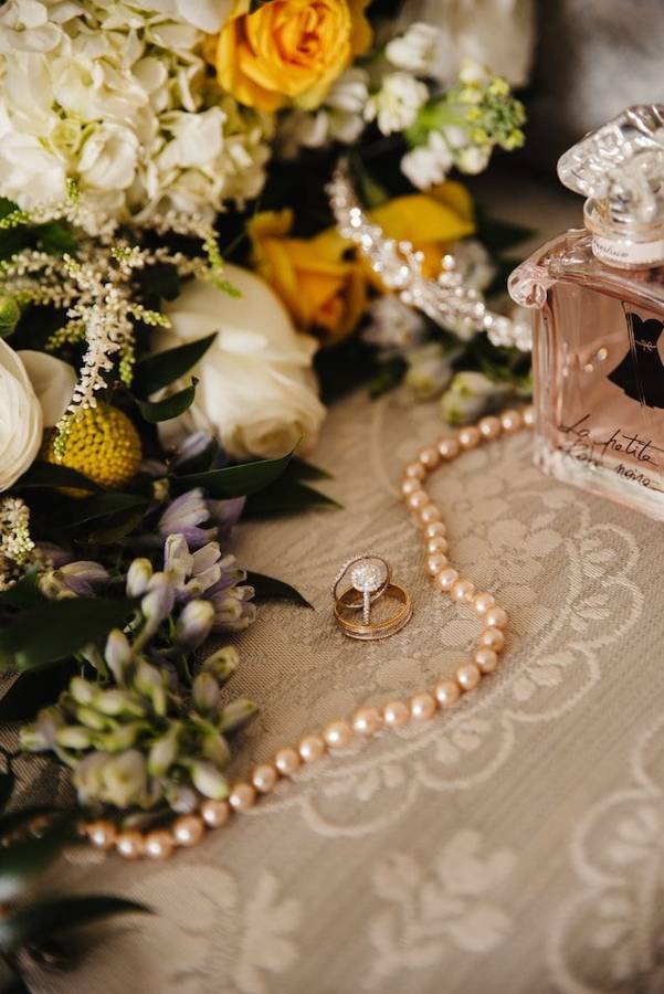 Wedding rings resting on lace beside a string of pearls, a perfume bottle and yellow and white flowers